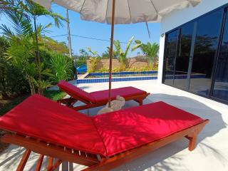 Two red sun loungers under a white umbrella with pool and lush greenery in the background