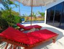 Two red sun loungers under a white umbrella with pool and lush greenery in the background