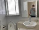 Modern bathroom with white fixtures, including a wall-mounted sink, toilet, and mirrored medicine cabinet, featuring venetian blinds on the window