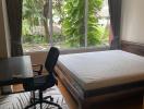 Bright bedroom with a large window overlooking greenery, featuring a study area and wooden flooring