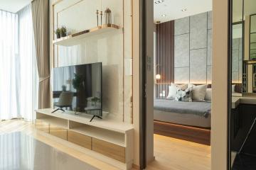 Modern living room with open space leading to bedroom, featuring large TV and reflective surfaces