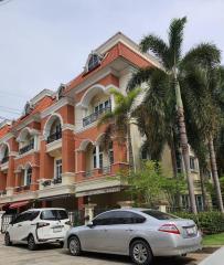 Elegant red brick apartment building with white trim and arched windows surrounded by green palm trees, with cars parked in front