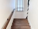 Wooden stairway leading up with white walls and a tall window with blinds