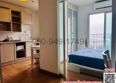 Compact bedroom with attached balcony, including a bed and work desk, adjacent to a small kitchen area
