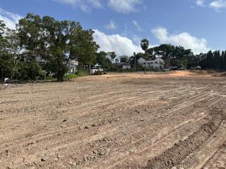 Large empty land plot ready for construction with nearby trees and residential structures in the background