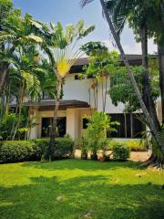 Lush green garden with tropical trees in front of a two-story house with large windows and a white facade