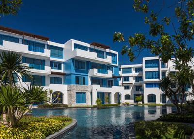 Modern apartment complex with blue and white facade surrounded by lush greenery and water feature