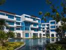 Modern apartment complex with blue and white facade surrounded by lush greenery and water feature