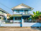 Two-story blue house with a fenced yard and tropical plants