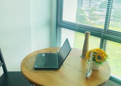 A neatly organized home office corner with a round table, laptop, and a vase of sunflowers by a window with blinds