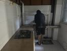 Person standing in a small, dimly lit kitchen with wooden cabinets and gas stove