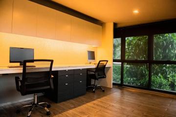 Modern home office with large desk, two chairs, and a view of greenery through a wall-sized window