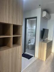 Compact apartment interior showing a wooden shelving unit, a glimpse of the bathroom, and a wall-mounted TV