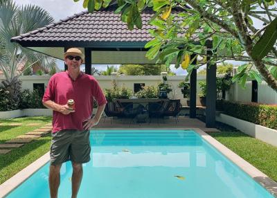 Man standing by the poolside with a drink in a backyard with lush greenery