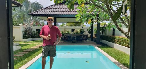 Man standing by the poolside with a drink in a backyard with lush greenery