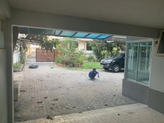 Spacious paved exterior with carport and greenery in the background