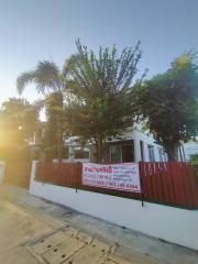 White single-story building with a green tree and fence, for sale sign in front