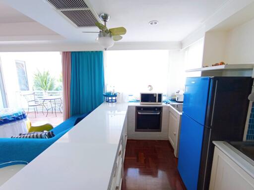 Modern kitchen with white countertops, blue refrigerator, and wooden flooring leading to a bright living area