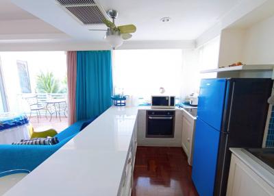 Modern kitchen with white countertops, blue refrigerator, and wooden flooring leading to a bright living area
