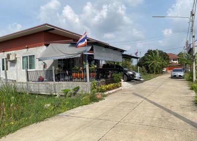 Single story residential home with red roof and extended awning in front, parked cars, and a flagpole with Thai flag