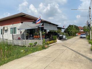 Single story residential home with red roof and extended awning in front, parked cars, and a flagpole with Thai flag