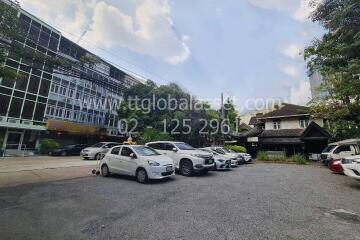 Parking area outside building with various cars and a tree-lined background