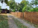 Brick fenced property with a gravel driveway leading to a wooden house surrounded by lush greenery