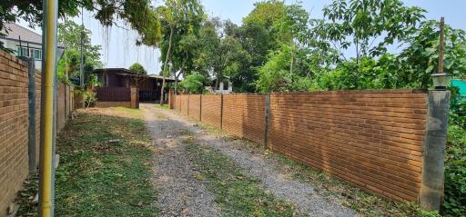 Brick fenced property with a gravel driveway leading to a wooden house surrounded by lush greenery