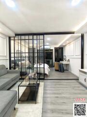 Modern apartment interior with open plan living space, comfortable grey couch, glass coffee table, and bedroom visible through glass partition