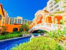 Luxurious resort-style building with a vibrant orange facade, arched bridge over a blue pool surrounded by lush greenery under a clear blue sky