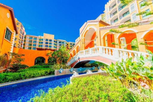 Luxurious resort-style building with a vibrant orange facade, arched bridge over a blue pool surrounded by lush greenery under a clear blue sky