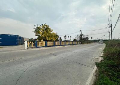 Paved street with a fenced property and blue shipping containers on the side under a cloudy sky