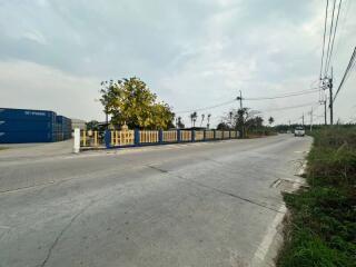 Paved street with a fenced property and blue shipping containers on the side under a cloudy sky