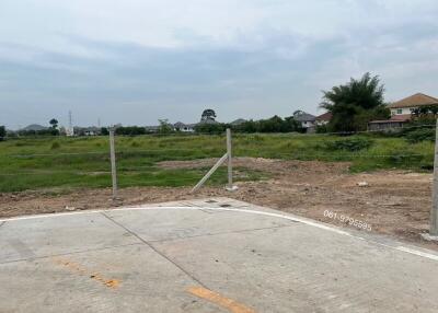 Vacant lot ready for construction with partial paved road in the foreground and scattered homes in the background under a cloudy sky