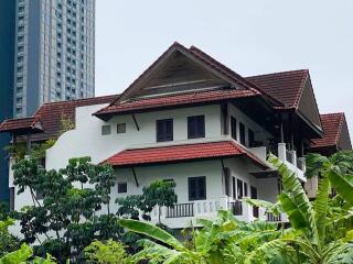 White multistory house with red roof surrounded by greenery near modern skyscrapers