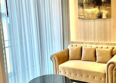 Elegant living room with natural light filtering through sheer curtains, featuring a plush beige sofa and a chic glass table