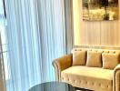 Elegant living room with natural light filtering through sheer curtains, featuring a plush beige sofa and a chic glass table