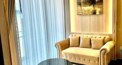 Elegant living room with natural light filtering through sheer curtains, featuring a plush beige sofa and a chic glass table