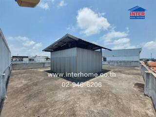 Exterior view of a small industrial warehouse surrounded by open space and other buildings under a clear sky