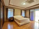 Spacious bedroom with wooden flooring, a large bed, wooden furniture, large windows with transparent drapes, and ceiling lighting