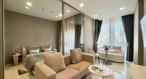 Modern open-plan living room connected with sleeping area featuring light brown couch, glass coffee table, and floor-to-ceiling windows with curtains