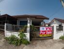 Single-story house with 'For Sale' sign, enclosed by a fence