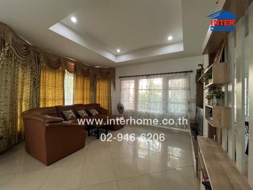 Spacious living room with brown sofa, large windows with drapes, tiled floor, and built-in shelving.