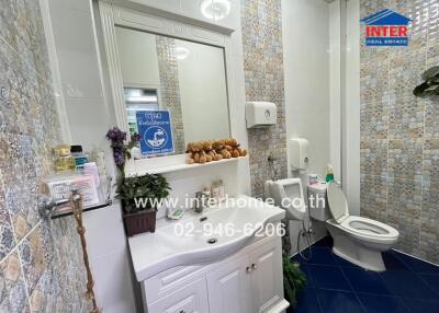 A modern, brightly lit bathroom with decorative tiles, a large mirror above a white vanity with a sink, toiletries, and plants. The bathroom also includes a toilet and a shower area.