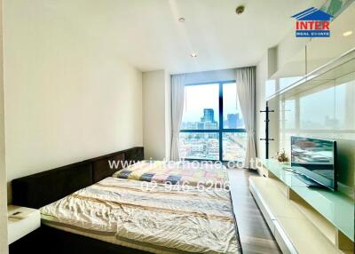 A spacious bedroom with a large window offering a city view, a double bed with bedding, a side table, and a wall-mounted TV with storage units.