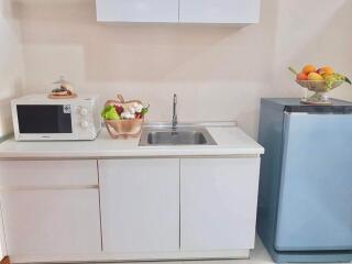 Compact kitchen area with white cabinets, a sink, a microwave, a small refrigerator, and a basket of fruits and vegetables.