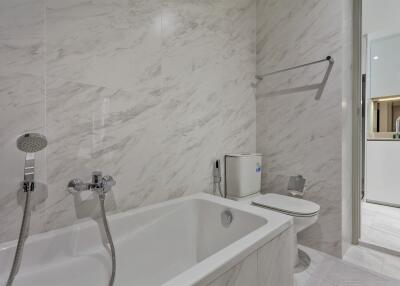 Modern bathroom with white marble walls, a bathtub, shower fixtures, toilet, and a glimpse into another room