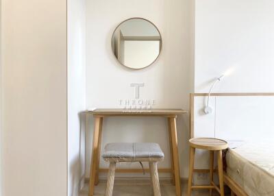 A small bedroom with minimalistic furniture including a bed, a stool at a wooden vanity table, circular mirror, and a bedside table