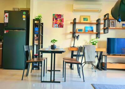 Modern living room and kitchen area with dining table and chairs, refrigerator, wall-mounted TV, shelves, and plants