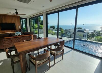 Spacious living area with large glass windows, wooden dining table, chairs, TV on the wall, and a view of the pool and sea.